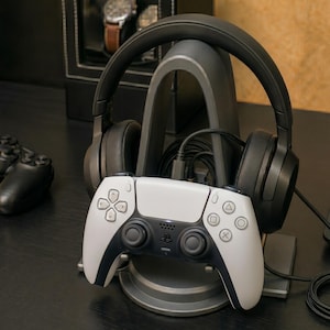 May include: A white and black PlayStation 5 controller rests on a gray stand, with black headphones and a charging cable. A black watch display case is in the background. The setup suggests a gaming or entertainment theme.