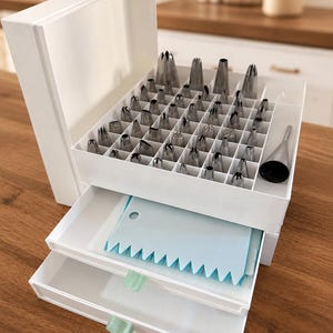 Op de afbeelding: Een witte opbergorganizer met meerdere niveaus die een verzameling zilverkleurige metalen spuitmondjes bevat in een bovenste vak met rasterindeling, met een lichtblauwe plastic deegschraper in de middelste lade en groene lipjes.