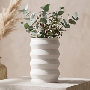 May include: A white, geometric vase filled with green and gray eucalyptus leaves and small seed pods. The vase has a ridged, zig-zag design and sits on a white surface. Dried pampas grass is visible in the background, adding a natural element to the composition.