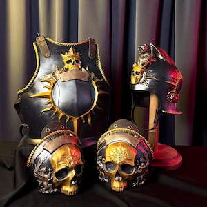 May include: Black and gold fantasy armor set, including a breastplate, two helmets, and two skull-shaped helmet fronts. The breastplate features a sunburst design and a skull emblem. The helmets are displayed on a wooden stand. The text "LARP SHOP KEEPER ONLINE ARMORY" is visible.