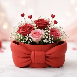Peut inclure: Un arrangement floral dans un récipient rouge en forme de nœud, composé de roses rouges et roses, de gypsophile et de cœurs décoratifs. L'ensemble est posé sur une surface blanche avec des confettis en forme de cœur.