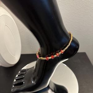 May include: A gold beaded anklet with red crystal beads. The anklet is displayed on a black mannequin foot, showcasing the jewelry's design and color. The anklet is a delicate piece of body jewelry.