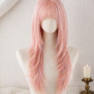 Puede incluir: Una peluca larga de color rosa pastel con capas y flequillo recto, exhibida en una cabeza de maniquí. El cabello presenta un color rosa claro suave con una textura lisa, cayendo más allá de los hombros en un corte en capas definido.