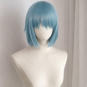 May include: A shoulder-length wig in a light blue colour with straight hair and bangs. The wig is displayed on a plain, off-white mannequin head and torso, showcasing the smooth texture and layered cut of the synthetic hair fibres in a neutral presentation.