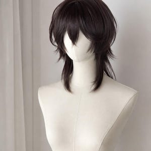 May include: A dark brown wig with a layered, shaggy cut featuring face-framing bangs and longer, textured strands at the back. The hair is styled with a voluminous crown and tapered ends, displayed on a smooth, off-white mannequin head and neck.