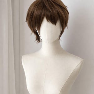 以下が含まれることがあります： 自然で無造作なスタイルに仕上げられた、レイヤー入りの短いブラウンのウィッグ。テクスチャーのある前髪とサイドの毛束が特徴です。ウィッグは滑らかなオフホワイトのマネキンの頭部と胴体に装着されており、ブラウンの色味とレイヤーカットが際立っています。