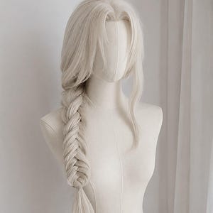 以下が含まれることがあります： 厚みのあるサイドフィッシュテール編みにスタイリングされた、長いプラチナブロンドのウィッグ。髪はセンターパートで、顔を縁取るルーズな毛束があり、ニュートラルな色調のマネキンの頭部と胴体のディスプレイフォームに装着されています。
