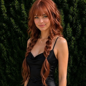 May include: A long, wavy, copper-red wig featuring straight-cut bangs and two loose, braided sections that fall over the shoulders. The hair has a vibrant, multi-tonal auburn hue with natural-looking highlights and a soft, layered texture throughout.