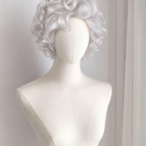May include: A short, curly wig in a light silver-gray color, styled with voluminous, soft curls that frame the head. The wig is displayed on a neutral-toned, fabric-covered mannequin bust, highlighting the texture and layered appearance of the hair.