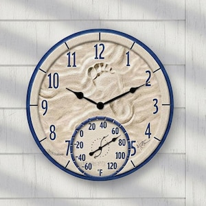 May include: A round wall clock with a beach theme. The clock face is beige with a footprint imprint and black hands. It has a blue border and a smaller thermometer below. The clock is mounted on a white wall.