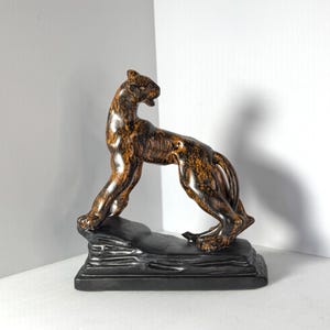 May include: A bronze-colored sculpture of a panther in a poised stance, atop a black rectangular base. The panther's body has a speckled pattern, with a dark brown and gold finish. The sculpture is approximately 12 inches long.