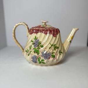 May include: Cream-colored ceramic teapot with a spiral design. The lid and body feature floral patterns with blue flowers and green leaves. The lid has a maroon border. The handle and spout are also cream-colored.
