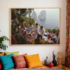 May include: A framed print shows a scenic outdoor dining scene. Four people are seated at a table laden with food, overlooking a body of water and rocky cliffs. The table is covered with a floral tablecloth. The print is displayed above a colourful rug and pillows.
