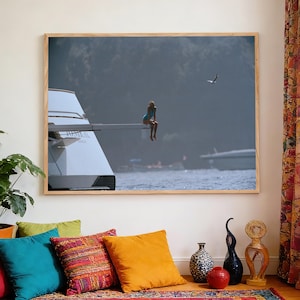 May include: A framed photograph shows a person in a blue swimsuit perched on a yacht's edge, gazing at the water. The scene is set against a backdrop of a tranquil seascape with a bird in flight. The room includes a vibrant rug and cushions.