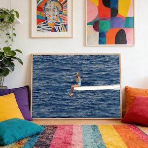 May include: A framed print of a person in a blue swimsuit sitting on a diving board over a vast, deep blue ocean. The image is framed by a light wood border and hangs on a white wall, with colorful abstract art above.