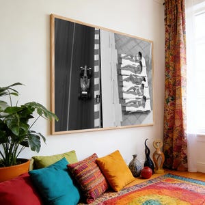 May include: Black and white framed photograph of a Formula 1 race car on a track, next to a group of people sunbathing on white lounge chairs. The photograph is displayed on a white wall above a colorful rug and pillows.