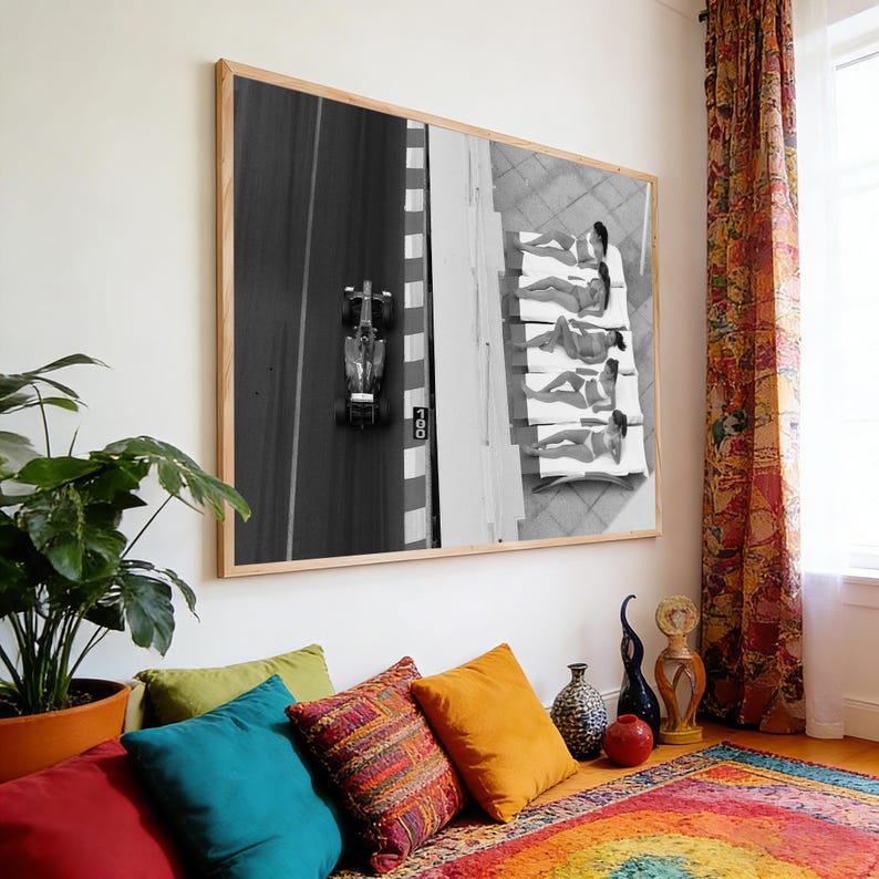 May include: Black and white framed photograph of a Formula 1 race car on a track, next to a group of people sunbathing on white lounge chairs. The photograph is displayed on a white wall above a colorful rug and pillows.