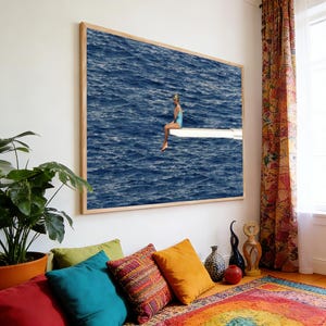May include: A framed print depicts a person in a blue swimsuit sitting on a diving board over a vast, dark blue ocean. The artwork is displayed on a white wall, with colorful pillows and a patterned rug in the foreground.
