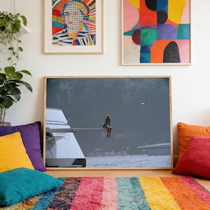 May include: A framed photograph depicts a person in a blue swimsuit sitting on the edge of a yacht, with a seagull in the sky. The image is set against a backdrop of water and a distant shoreline. The room features colorful pillows and a striped rug.