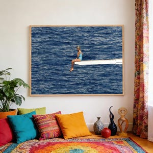 May include: A framed print depicts a person in a blue swimsuit sitting on a diving board over a vast, dark blue ocean. The artwork is displayed above a colorful rug and pillows, with a potted plant and decorative vases nearby.
