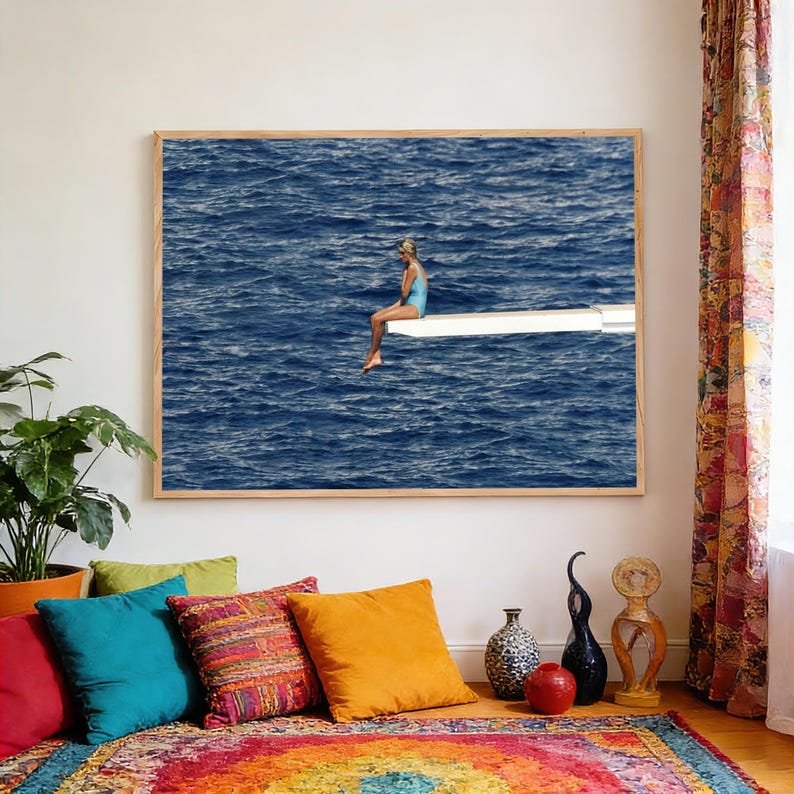 May include: A framed print depicts a person in a blue swimsuit sitting on a diving board over a vast, dark blue ocean. The artwork is displayed above a colorful rug and pillows, with a potted plant and decorative vases nearby.