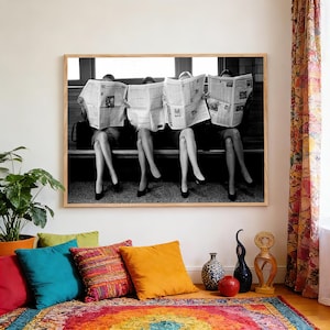 May include: A monochrome framed print showing four people reading newspapers, focusing on their legs and feet in black heels. The print is above a rug and colorful cushions. The image is in a room.