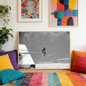 May include: Black and white framed photograph of a person sitting on a diving board over water. The image is in a room with colorful abstract art and pillows. A multi-colored striped rug is in the foreground.