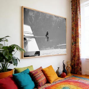 May include: Black and white framed photograph of a person sitting on the edge of a yacht, overlooking the water. The image is mounted on a light wood frame and hangs above a colorful rug and pillows.