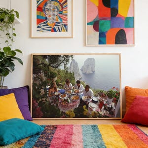 Puede incluir: Fotograf&iacute;a enmarcada que representa una escena de comedor al aire libre con vistas al mar. La mesa est&aacute; puesta con comida y rodeada de gente. Cojines coloridos y una alfombra a rayas en primer plano, con arte abstracto en la pared.