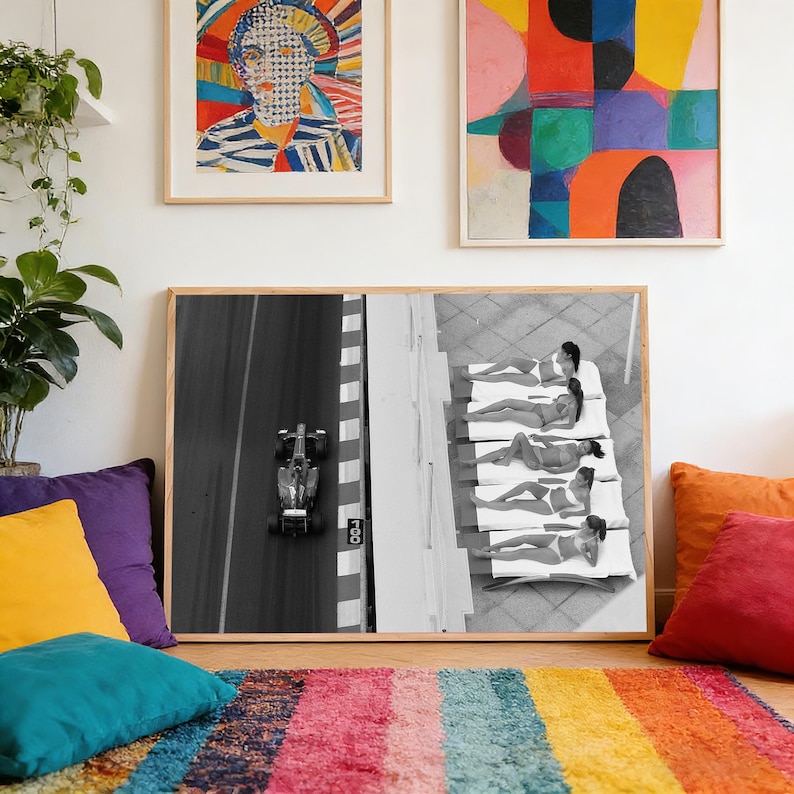 May include: Black and white framed print featuring a Formula 1 race car on a track and people sunbathing by a pool. The print is displayed with colorful abstract art and a portrait. A rainbow rug and pillows add color.