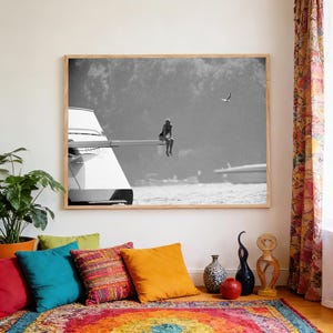May include: Black and white framed photograph of a person sitting on the edge of a boat. The image is in a wooden frame and is displayed in a room with colorful pillows and a patterned rug. A bird flies in the background.