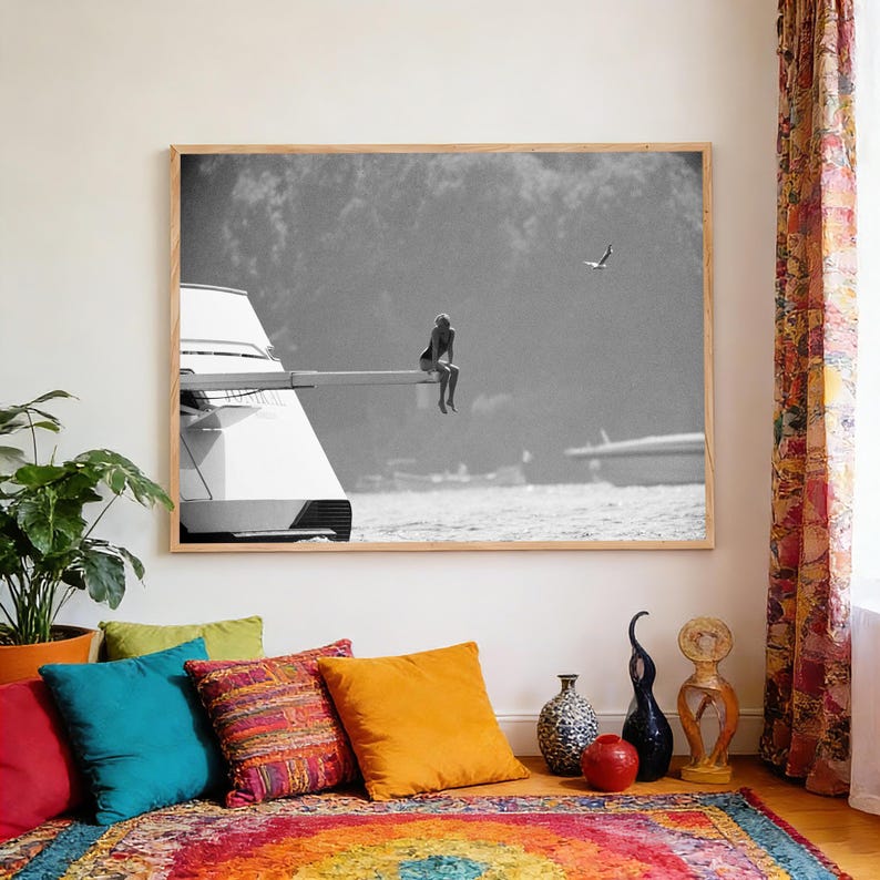May include: Black and white framed photograph of a person sitting on the edge of a boat. The image is in a wooden frame and is displayed in a room with colorful pillows and a patterned rug. A bird flies in the background.