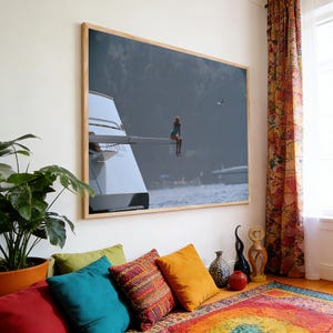 May include: A framed photograph depicts a person sitting on the edge of a yacht's wing, overlooking a body of water. The image is framed in a light wood frame and hangs on a white wall. The room features colorful pillows, a patterned rug, and a potted plant.
