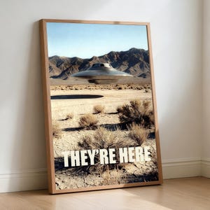 Könnte beinhalten: Ein gerahmter Druck, der eine Wüstenlandschaft mit einer metallischen fliegenden Untertasse zeigt, die über der Landschaft schwebt. Der Druck enthält den Text "THEY'RE HERE" in fetten weißen Buchstaben vor dem Hintergrund von Bergen und trockenem Gelände.