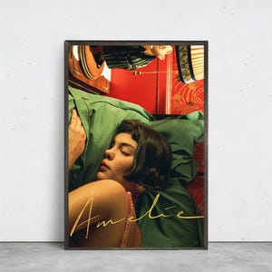 以下が含まれることがあります： 緑色の枕で休んでいる女性が描かれた額入りのプリント。金色の筆記体で「Amelie」の文字が書かれています。背景には赤と模様の要素があり、ヴィンテージな美学を作り出しています。プリントは濃い木製のフレームに収められています。