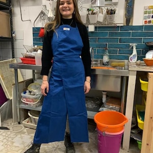 Może przedstawiać: Niebieski fartuch z małą białą metką jest noszony na czarnej koszuli z długim rękawem. Fartuch zakrywa nogi i jest noszony z czarnymi butami. Tło obejmuje zlewozmywak ze stali nierdzewnej, półki i różne przedmioty kuchenne.