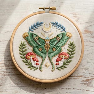Op de afbeelding: Geborduurd kunstwerk in een houten ring. Het ontwerp toont een groene mot met gedetailleerde vleugels, omringd door bladeren, paddenstoelen, een maan en een ster. De borduursels zijn op een crèmekleurige stof.