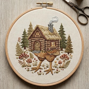 May include: Embroidered artwork in a wooden hoop features a whimsical cabin scene. The cabin has a thatched roof and a chimney with smoke. A creature with chicken legs supports the cabin. Mushrooms and foliage surround the cabin.