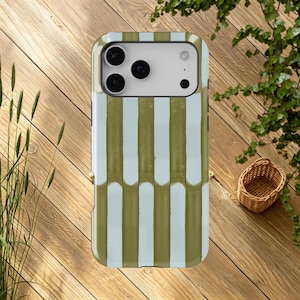 May include: A smartphone case featuring a vertical striped design in olive green and pale blue. The case is placed on a wooden surface, with a small woven basket and foliage in the background.