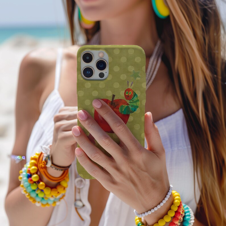 Puede incluir: Funda de tel&eacute;fono verde con una ilustraci&oacute;n de una manzana roja y una oruga colorida. La funda tiene un patr&oacute;n de lunares y se sostiene frente a un fondo de playa.