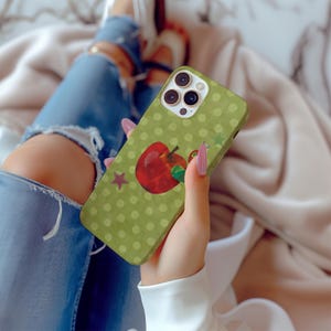 Puede incluir: Una funda de tel&eacute;fono verde con un dise&ntilde;o de lunares y una ilustraci&oacute;n de una manzana roja. Tambi&eacute;n hay una estrella roja en la funda. La funda del tel&eacute;fono la sostiene una persona que lleva vaqueros azules.
