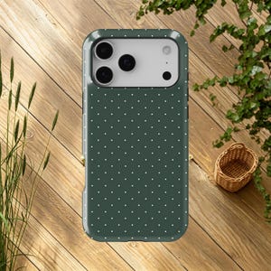 May include: A dark green phone case with a white polka dot pattern. The case has a glossy finish and a camera cutout. The phone case is on a wooden surface with a small woven basket and greenery in the background.