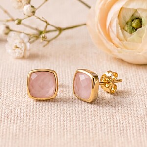 Puede incluir: Un par de pendientes de botón con piedras facetadas de color rosa pálido en forma cuadrada, engastadas en metal dorado pulido. Cada pendiente cuenta con un cierre de presión seguro, mostrando un diseño geométrico limpio y acabado translúcido.