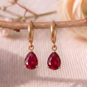 Pode incluir: Um par de brincos de argola dourados com pedras preciosas vermelhas pendentes em forma de lágrima. Cada brinco consiste num aro fino de ouro ligado a uma pedra vermelha profunda facetada em forma de pera que reflete a luz intensamente.