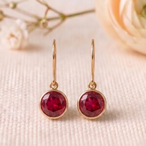 May include: A pair of gold drop earrings featuring round, faceted, deep red stones set in a simple metal bezel. The earrings hang from thin, curved gold wires, showcasing a polished finish and a classic, symmetrical design that catches the light.