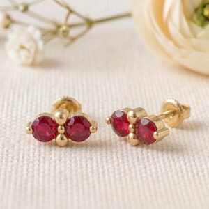 May include: A pair of gold stud earrings, each featuring two round, faceted, deep red stones set horizontally side-by-side. The stones are separated by a small gold bead, and the earrings include gold post backs for secure attachment to the ear.