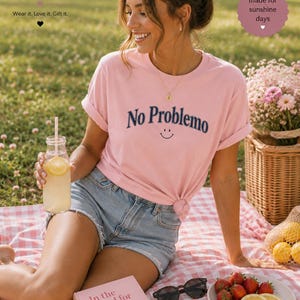 May include: A pink t-shirt with the words "No Problemo" and a smiley face, paired with denim shorts. The image includes a picnic setting with a book, sunglasses, strawberries, and a drink. The Boffy Boutique logo is visible.