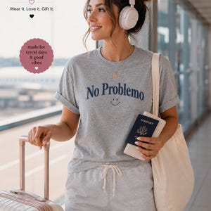 May include: A woman wearing a gray t-shirt with "No Problemo" and a smiley face graphic, paired with gray shorts. She holds a passport and a tote bag, and is near a suitcase. The image includes the text "Boffy Boutique".