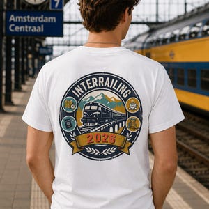 May include: A white short-sleeved t-shirt featuring a circular graphic on the back. The design depicts a train on a bridge, mountains, and travel icons including a map, passport, coffee cup, and backpack, with the text "INTERRAILING 2026".