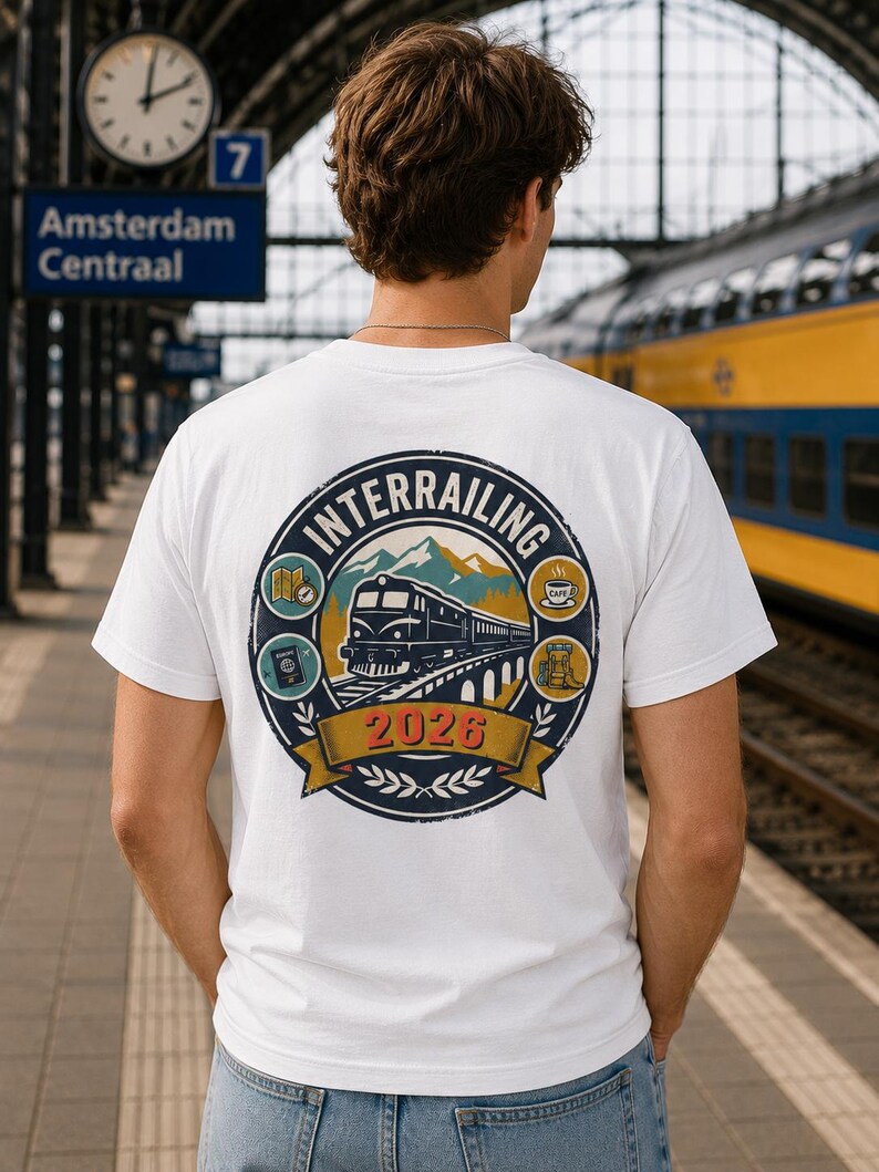 May include: A white short-sleeved t-shirt featuring a circular graphic on the back. The design depicts a train on a bridge, mountains, and travel icons including a map, passport, coffee cup, and backpack, with the text "INTERRAILING 2026".